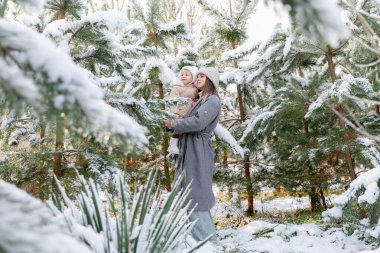 Anne karlı bir çam ormanında gülen bebeğini kucağında tutuyor. Dallardan kar yağarken neşeli bir kış anı.