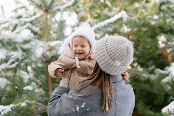 Anne gülen bebeğini karlı bir çam ormanında kaldırıyor. Doğal ışık ve kalın giysilerle neşeli aile kış anı.