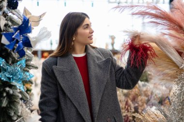 Kadın mağazadan şenlikli dekorasyon alışverişi yapıyor. Şık kış kıyafetleri ve tatil süsleri sıcak, mevsimsel bir atmosfer yaratır..