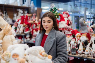 Raflardaki dekoratif yumuşak figürlere bakan genç bir kadın. Çeşitli konforlu eşyalar ve parlak arka plan ışıklarıyla kapalı perakende sahne.