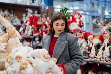 Raflardaki dekoratif yumuşak figürlere bakan genç bir kadın. Çeşitli konforlu eşyalar ve parlak arka plan ışıklarıyla kapalı perakende sahne.
