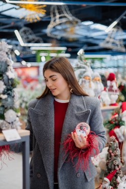 Genç bir kadın dekoratif bir kar küresini incelerken mevsimlik dcor ile dolu bir mağazanın içindeki raflardaki eşyaları inceliyor..