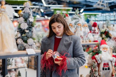 Genç bir kadın dekoratif bir kar küresini incelerken mevsimlik dcor ile dolu bir mağazanın içindeki raflardaki eşyaları inceliyor..