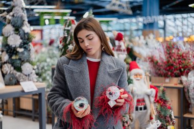 Mevsimlik dcor ile dolu bir mağaza koridorunda dururken iki dekoratif eşyayı karşılaştıran genç bir kadın hangi ürünü seçeceğini dikkatlice değerlendiriyor..