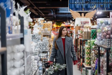 Genç bir kadın dekoratif eşyalarla dolu bir koridorda yürürken elinde yeşil bir alışveriş sepetiyle etrafındaki raflara bakıyor..