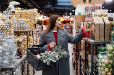 Genç bir kadın dekoratif eşyalarla dolu bir koridorda yürürken elinde yeşil bir alışveriş sepetiyle etrafındaki raflara bakıyor..