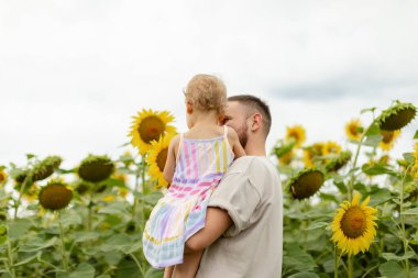 Babam bir ayçiçeği tarlasında yürürken kucağında bir bebekle parlak sarı ayçiçeklerine bakıyordu. Sıcak yaz sahnesi, aile bağlantısı, doğa ve kırsal alan.