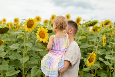 Babam bir ayçiçeği tarlasında yürürken kucağında bir bebekle parlak sarı ayçiçeklerine bakıyordu. Sıcak yaz sahnesi, aile bağlantısı, doğa ve kırsal alan.