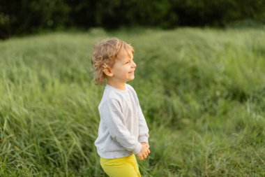 Güneşli bir günde uzun yeşil çimenlerin üzerinde gülümseyen bir bebek. Açık havada sıcak ışık, neşeli ifade ve doğal çocukluk enerjisiyle.