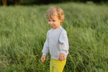 Uzun otların arasında yürüyen neşeli çocuk, yumuşak doğal ışıkta komik bir surat yapıyor. Açık havada gerçek çocukluk kişiliği ve hareketini gösterme anı.