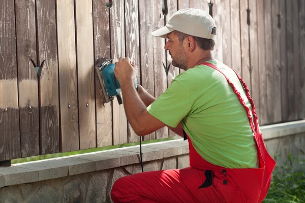 Trabalhador lixando pintura velha de uma cerca de madeira — Fotografia de Stock