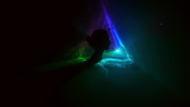 暗い中で移動するさまざまな色の光の背景に若い女の子のイメージ.煙は女の子の頭で三角形を埋めます.プロジェクター. — ストック動画
