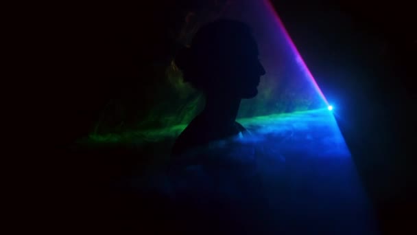 Зображення молодої дівчини на світловому фоні різних кольорів, що рухаються в темряві. Дим заповнює трикутник з головою дівчат. — стокове відео