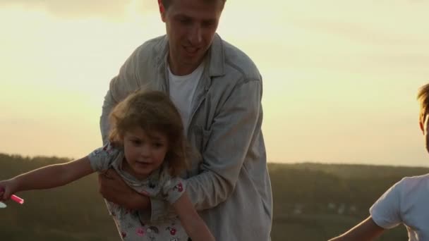 Familie vakantie in de natuur. Papa houdt een dochtertje vast en zijn zoon rent rond. Man Twists zijn dochter alsof ze vliegen. — Stockvideo