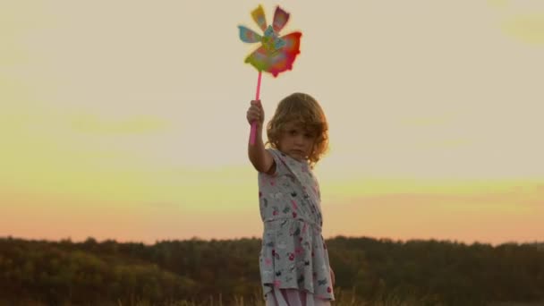 Bambina in piedi sulla natura al tramonto. Ragazza che tiene giocattolo mulino a vento che si muove nel vento — Video Stock