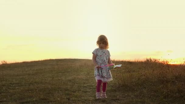 日落时站在自然之上的小女孩。在风中摇曳着玩具风车的女孩. — 图库视频影像