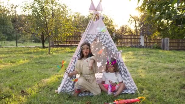 Kinderurlaub. Zwei hübsche Mädchen in Indianerkostümen sitzen im Zelt im Garten. — Stockvideo