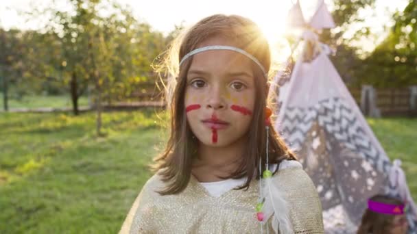 Het gezicht van een klein meisje gekleed als een indiaan. Meisje met donker haar en beschilderd gezicht. Veren op haar hoofd. — Stockvideo