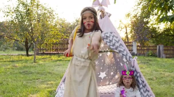 Barnens semester. Två söta flickor i kostymer av indianer sitter i tält i trädgården. — Stockvideo