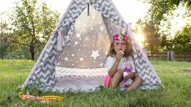 Mała kręcona dziewczynka siedząca w namiocie w ogrodzie i jedząca pianki. Jest udekorowana jak Indianka. Cute little girl z piórami na głowie i malowane twarz. — Wideo stockowe
