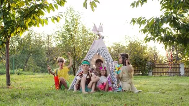 Fünf Kinder spielen Indianer. Drei Jungen und zwei Mädchen sitzen mit aufgemalten Gesichtern und Pfeilen in den Händen im Zelt draußen.. — Stockvideo
