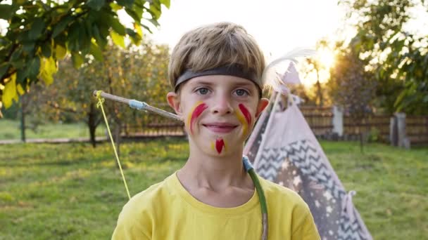 Un chico guapo a imagen de un indio. Sostiene una flecha y grita. Lindo chico con cara pintada y diadema. Detrás de él tienda en el jardín. Niño jugando indio líder. — Vídeos de Stock