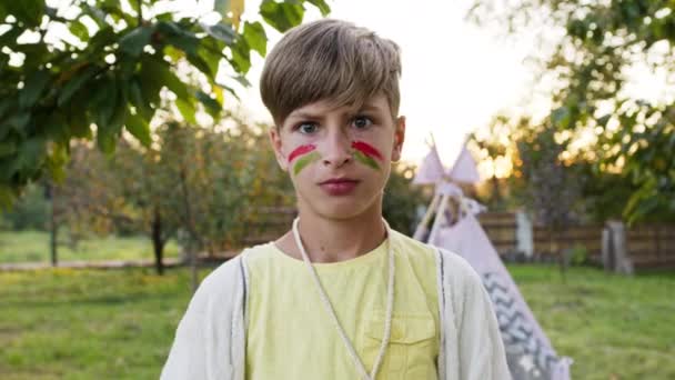 Niño con la cara pintada. Es serio y en la imagen de un indio. Detrás de él tienda en el jardín. — Vídeos de Stock