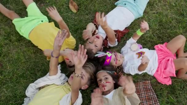 Fünf Kinder liegen auf dem Rasen, die Köpfe stehen einander gegenüber. Schießen von oben. Kinder in den Bildern von Indianern mit gemalten Gesichtern. — Stockvideo
