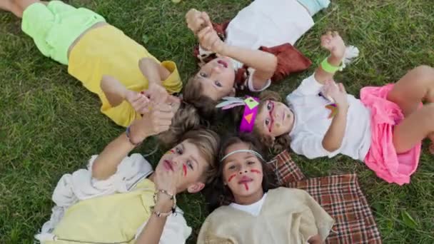 Cinco niños yacen en la hierba con la cabeza mirando el uno al otro. Disparando desde arriba. Niños en las imágenes de indios con caras pintadas. — Vídeos de Stock