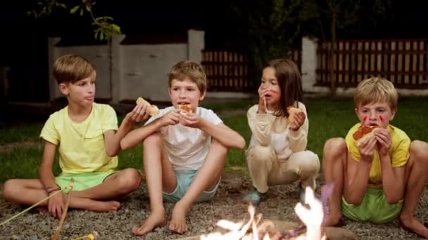 Gyerekek ülnek a tűz mellett az utcán és esznek. Belefáradtak a szórakozásba. A gyerekek indiánosat játszottak. Arcokat festettek. Jó estét.. — Stock videók