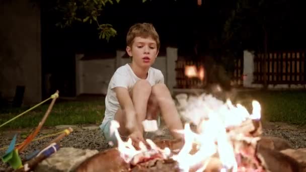Sömnig pojke sitter vid elden och håller något på pinne och steker på elden. — Stockvideo