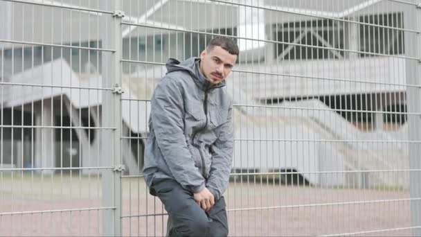 Retrato de un joven serio en la calle. Hombre guapo en chaqueta gris con cuello alto. Chico está parado cerca de valla de metal. — Vídeos de Stock