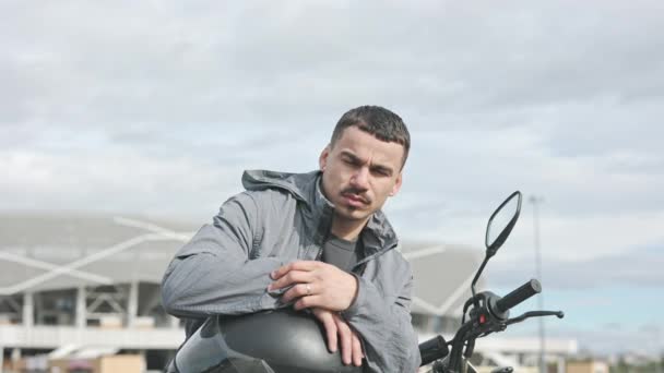 Joven sentado en motocicleta. Está sosteniendo el casco. lindo oscuro pelo chico con bigote en la calle. — Vídeos de Stock