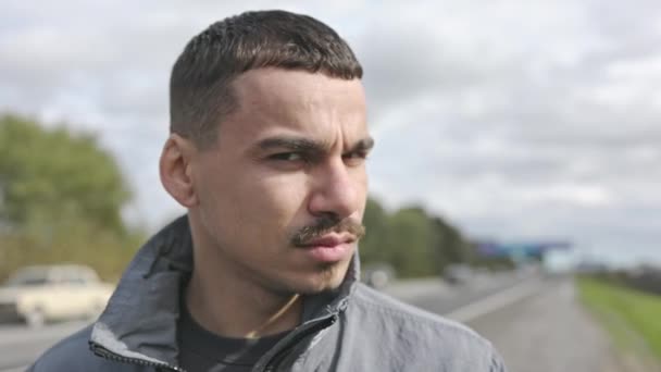 Portret van een serieuze jongeman op straat. Bruut beeld van de man op straat. Achter hem De weg. — Stockvideo