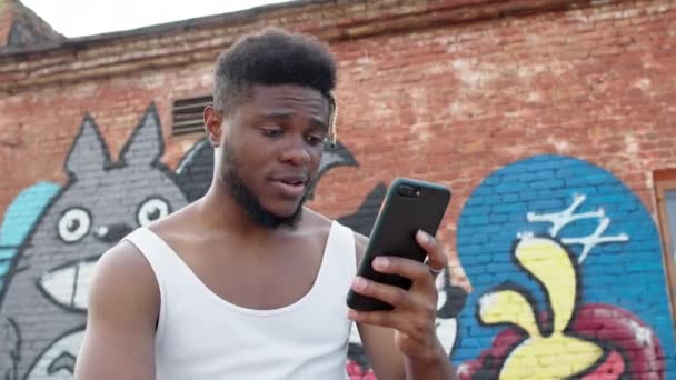 Jonge zwarte man met mobiele telefoon in zijn handen. Man zit op straat met geschilderde muren van huizen.. — Stockvideo