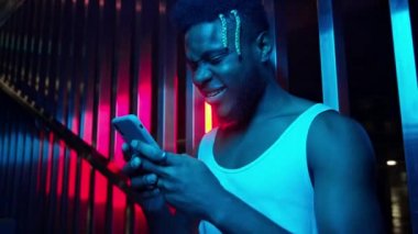 Elinde telefon olan genç siyahi adam. Yeraltı otoparkının yanındaki merdivenlerde duruyor ve gülümsüyor. Açık Mavi ve Pembe Renkler.