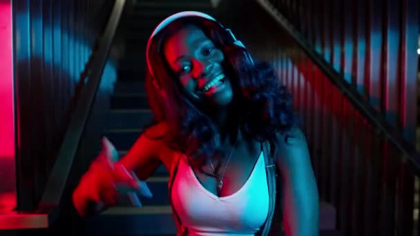 Portrait Of Black Young Girl In Headphones. She Stands On The Background Of The Stairs. Lighting In Different Colors. — Stock Video