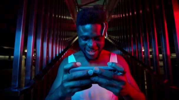 Jonge zwarte man met telefoon in zijn handen. Hij staat op de trappen in de buurt van de ondergrondse parkeerplaats en lacht. Lichtblauwe en roze kleuren. — Stockvideo