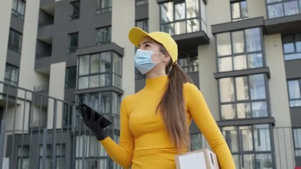 Fiatal lány a szállító cégtől dobozt hord. Orvosi maszkot és kesztyűt visel, hogy megvédje a vírusfertőzéstől. A kezében van a telefonja. A lány néz ki az ablakon a magas emeleti épület. — Stock videók