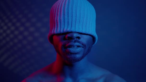 Черный парень в комнате с голубым светом. На его голове шляпа, которая закрывает его глаза. — стоковое видео