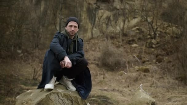 Stijlvolle jongeman in de natuur. Aardige vent in lange jas en pet op Stone. — Stockvideo