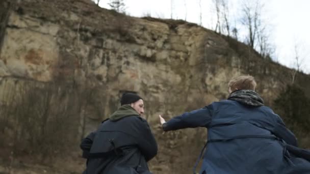 Twee jongemannen rennen vooruit. Jongens in regenjassen op Rock achtergrond. Uitzicht vanaf de achterkant. — Stockvideo