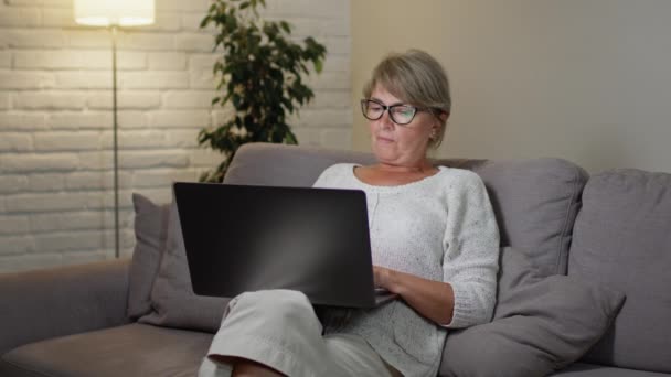 Kobieta używa laptopa. Blondynka w okularach z ładnym wyglądem spoczywa na kanapie wieczorem. Przegląda coś w Internecie lub komunikuje się przez portale społecznościowe. — Wideo stockowe