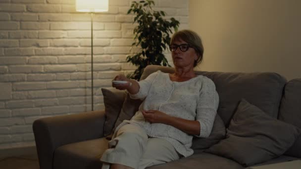 Gyönyörű nő ül a kanapén a lakásában. Ő tartja a távirányítót a TV-ből, és csatornát vált.. — Stock videók