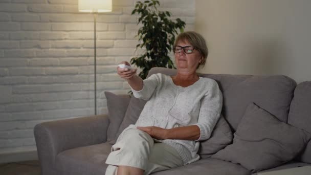 Middeleeuwse vrouw zit op de bank in haar appartement. Ze houdt de afstandsbediening van de TV en wisselt van kanaal. Licht in de kamer verandert. — Stockvideo