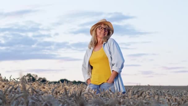 Lekfull bild av blond i glasögon och hatt. Kvinnan står på vetefält och ler. Hon håller händerna i byxorna Fickor. — Stockvideo