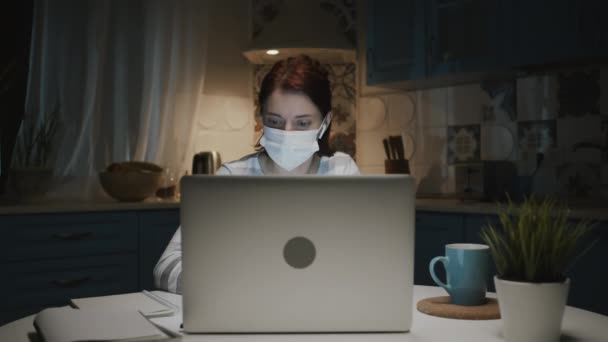 Rapariga na cozinha com laptop. Mulher em máscara médica faz notas no bloco de notas. — Vídeo de Stock