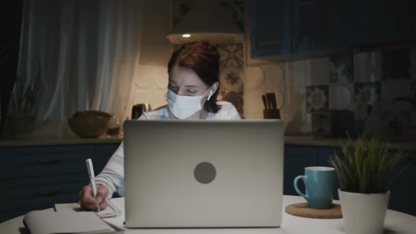 Giovane ragazza in cucina con computer portatile. Donna in maschera medica prende appunti nel blocco note. — Video Stock