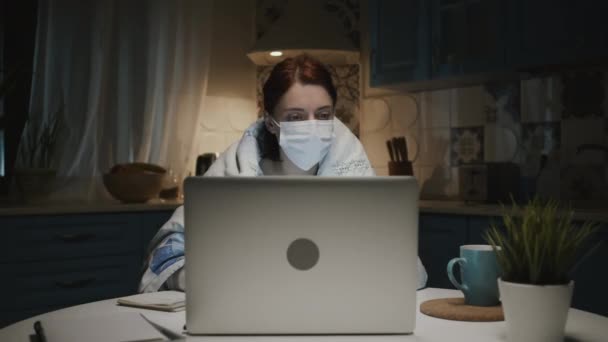 Ung flicka i köket med laptop. Ung kvinna arbetar på distans hemma på grund av en pandemi. Kvinna i medicinsk mask. — Stockvideo