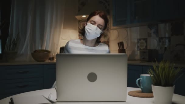 Ung flicka i köket med laptop. Ung kvinna arbetar på distans hemma på grund av en pandemi. Kvinna i medicinsk mask. — Stockvideo